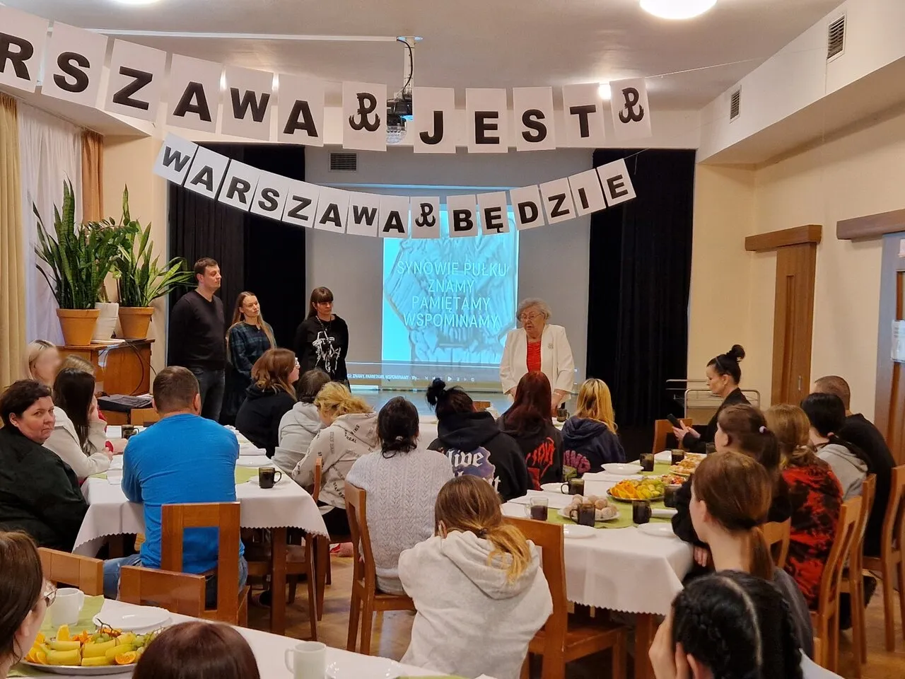 Młodzież siedzi w sali i słucha opowiadania starszej kobiety