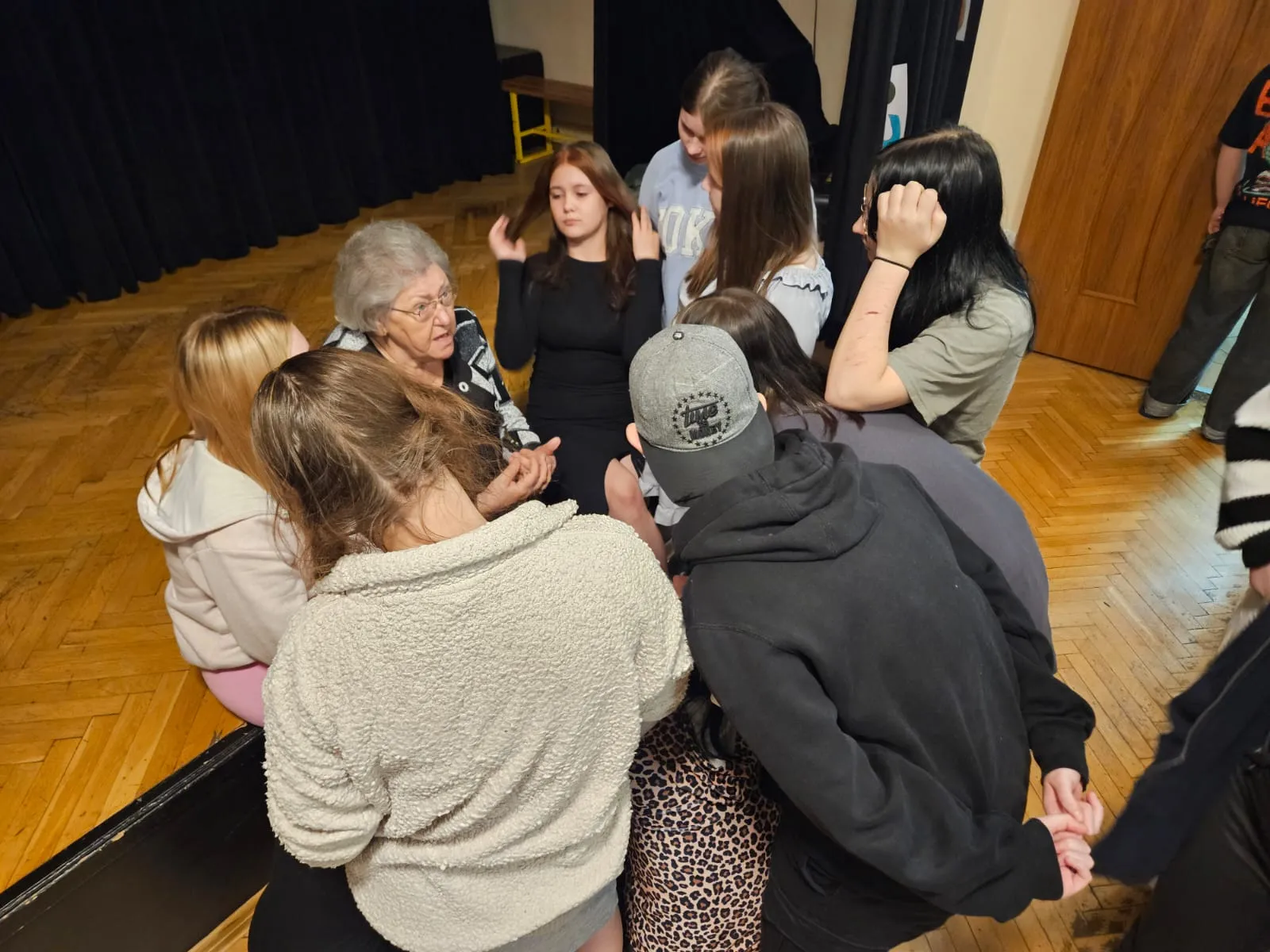 Młodzież siedzi w sali i słucha opowiadania starszej kobiety