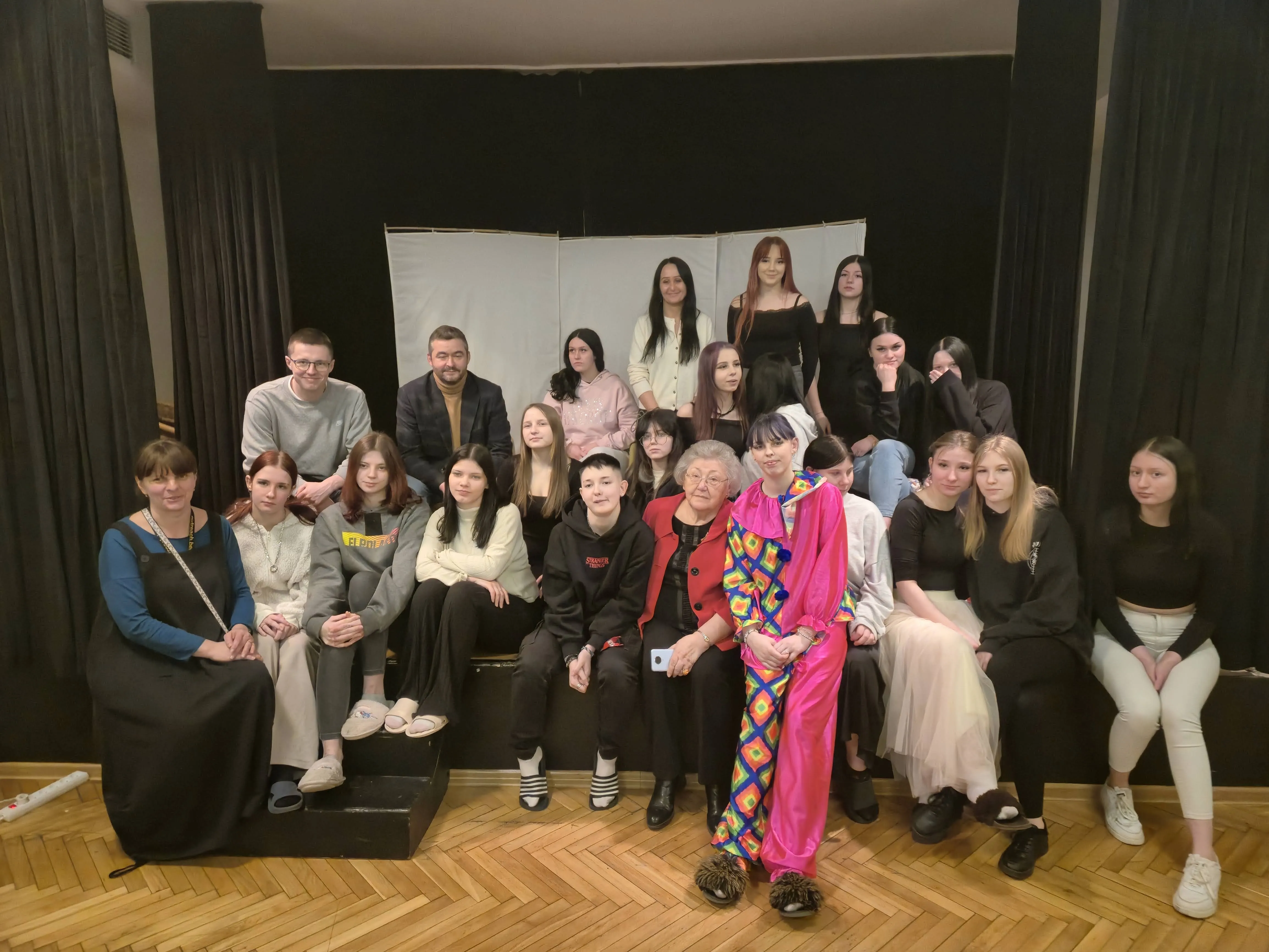Grupa młodzieży siedzi na scenie ze starszą panią